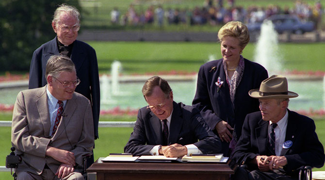 President George H.W. Bush sitting at the South Lawn of the White House putting pen to paper on the Americans with Disabilities Act surrounded by 4 individuals